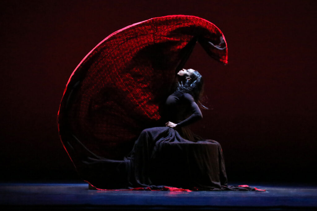 On a darkened stage, Leslie Andrea Williams is seated in profile, face upturned as a wave of red fabric flies behind her and overhead. Her downstage arm is pulled tight to her side, elbow drawn back behind her ribs.