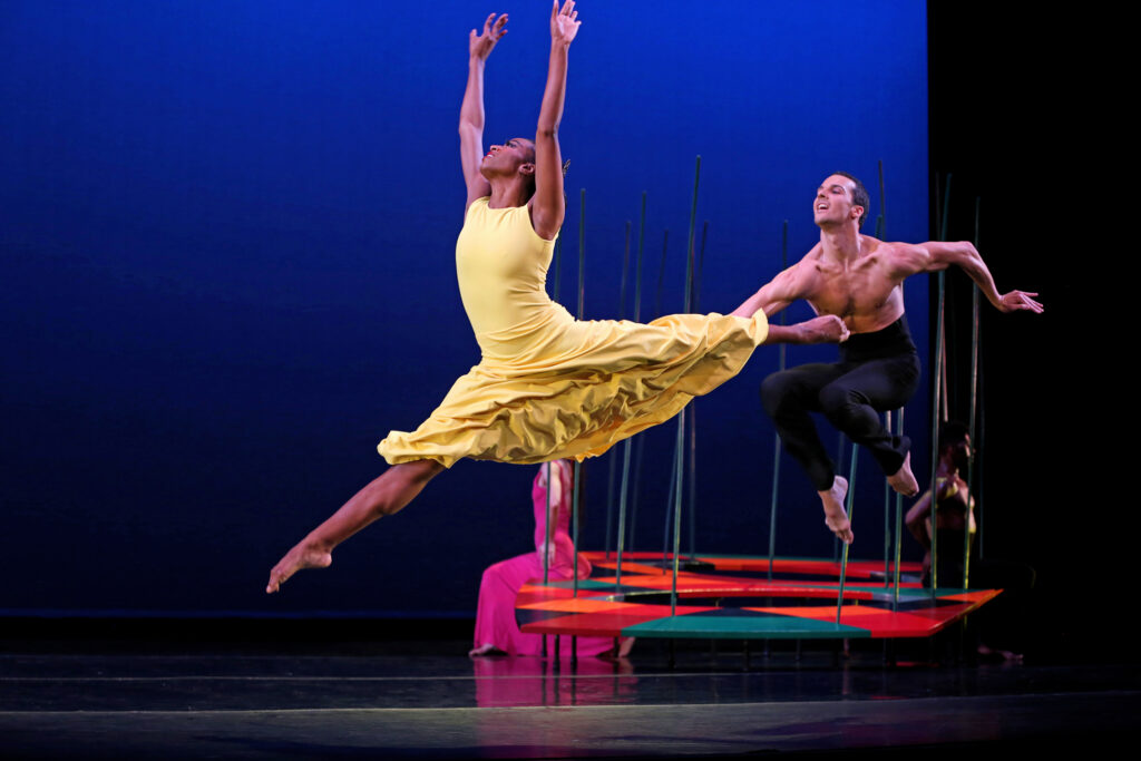 Leslie Andrea Williams is captured mid-leap, her arms pressing overhead and chin tipped up. She is barefoot and wears a sleeveless yellow dress. Behind her, a male dancer jumps, tucking both knees up to his chest and looking out toward her. A series of colorfully patterned platforms with poles sticking up out of them are arrayed upstage.