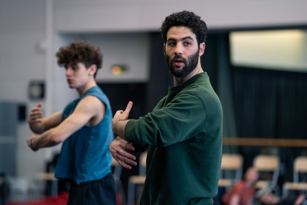 Or Schraiber looks forward over his shoulder, demonstrating a gesture as though bringing an invisible person in for a hug. A dancer in the rehearsal studio behind him imitates the motion.