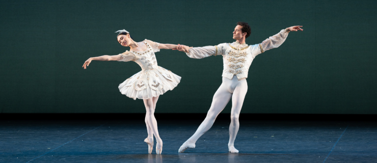 Cory Stearns in tendu effacé front, supporting knee in plié, as he extends a hand to support Hee Seo. She balances in sous-sus, one hand in his as she leans away from him. Their outside arms reach away from each other. She wears a classical tutu, pink tights, and pointe shoes, while he is in a tunic, white tights, and ballet slippers, both in matching white with gold accents.