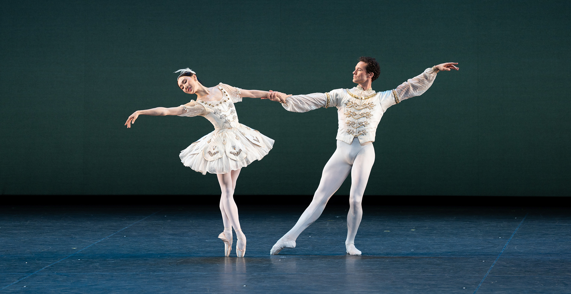 Cory Stearns in tendu effacé front, supporting knee in plié, as he extends a hand to support Hee Seo. She balances in sous-sus, one hand in his as she leans away from him. Their outside arms reach away from each other. She wears a classical tutu, pink tights, and pointe shoes, while he is in a tunic, white tights, and ballet slippers, both in matching white with gold accents.