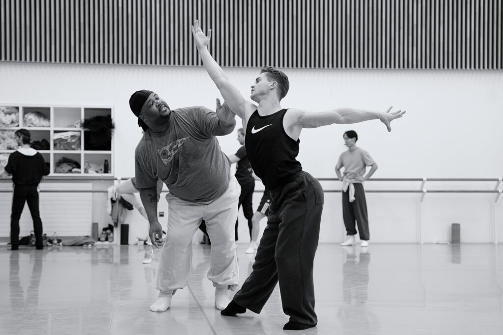 In a rehearsal studio, Kameron N. Saunders adjusts the arm of a dancer as it is raised forward. The dancer sits into his back hip, back arching.