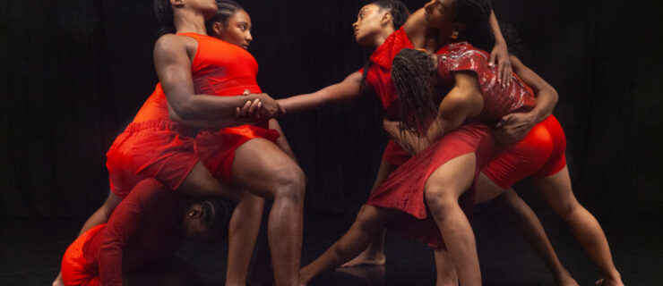 Two clusters of three dancers in red face each other. One stretches an arm across the space between them to clasp the hand of another, whose eyes are closed as they lean back into the dancer bracing them. Limbs are interwoven and weight shared.