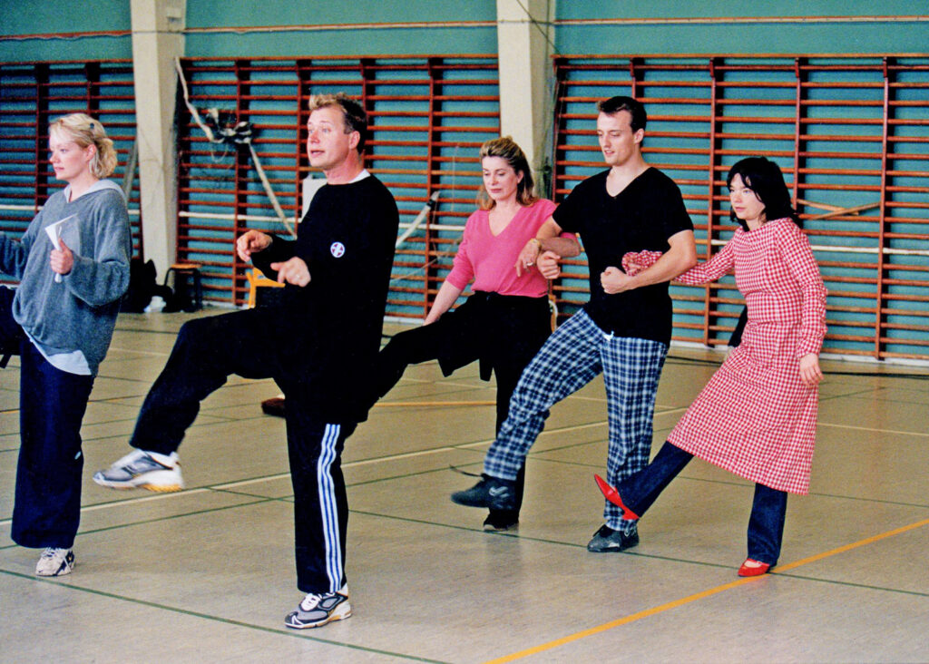 Vincent Paterson teaching cast members steps. They link arms as a trio and kick their right leg forward. 