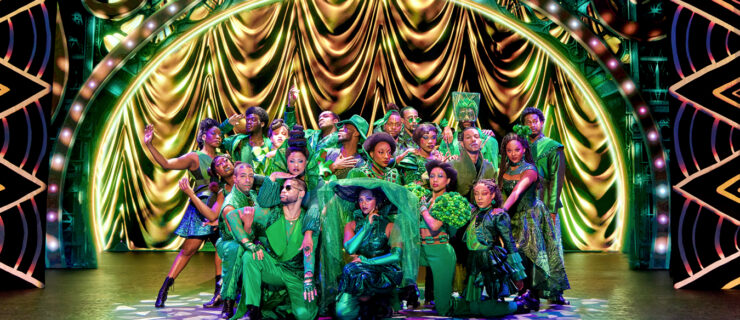 The cast of The Wiz strike a pose on stage. They wear a variety of green costumes with large hats, sparkly jackets, and patterned skirts.