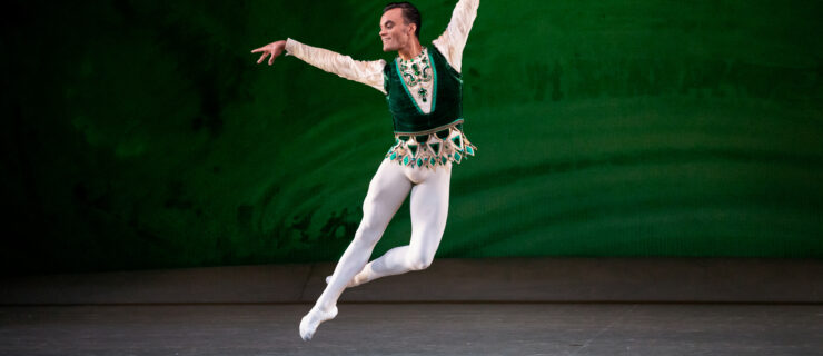 Taylor Stanley is captured midair in a petit jeté, one foot tucked into coupé back. Their arms are in allongé. They are costume in white tights and ballet slippers with an emerald green tunic vest over a long sleeved off-white shirt.