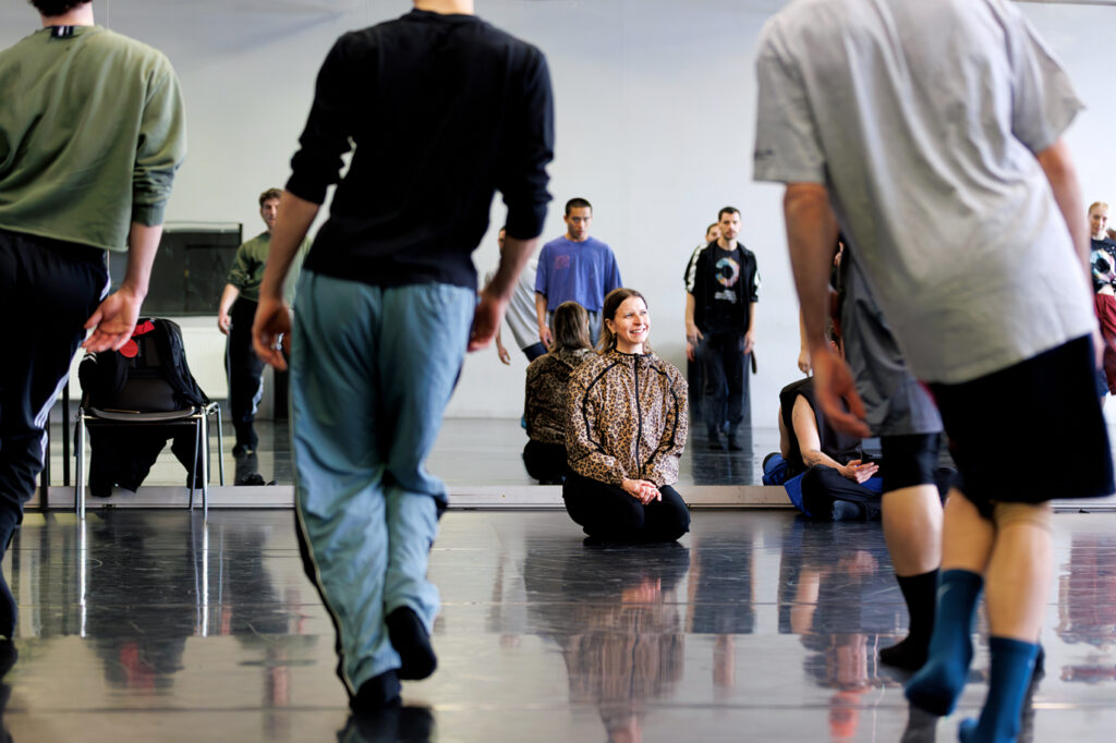 Aszure Barton kneels at the front of a rehearsal studio, smiling as dancers rehearse.