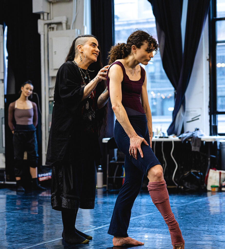 Sonya Tayeh stands behind Melanie Moore in a studio, fingertips on her shoulders. Moore props one foot on forced arch, head stretching forward as she hunches her shoulders slightly.