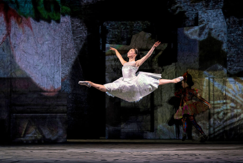 Madeline Woo at the height of a lofty grande jeté with arms in allongé. She smiles radiantly, chin upraised. She is costumed in a flowing silver gown with pointe shoes painted and bejeweled to match.