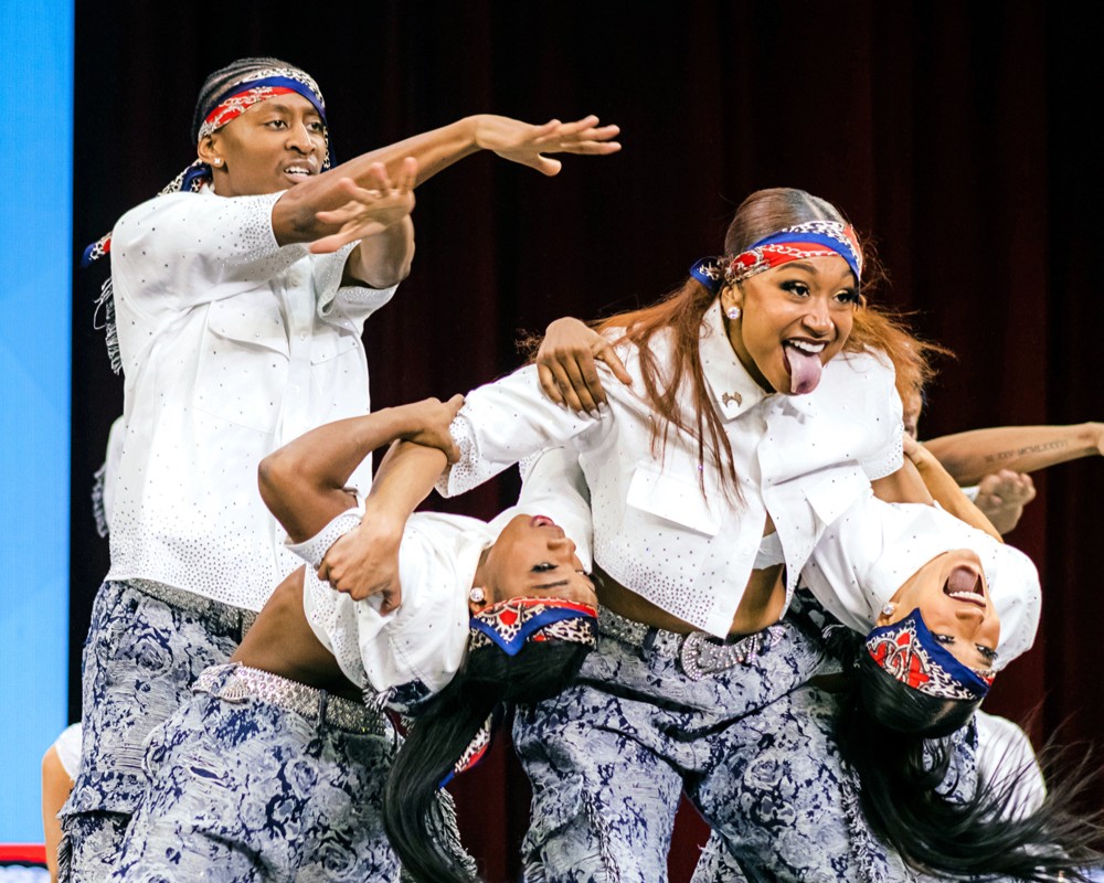 Maya Ramey sticks her tongue out while supporting two other dancers as they bend back and look towards the audience. The team wears patterned pants, white button down shirts, and bandanas.