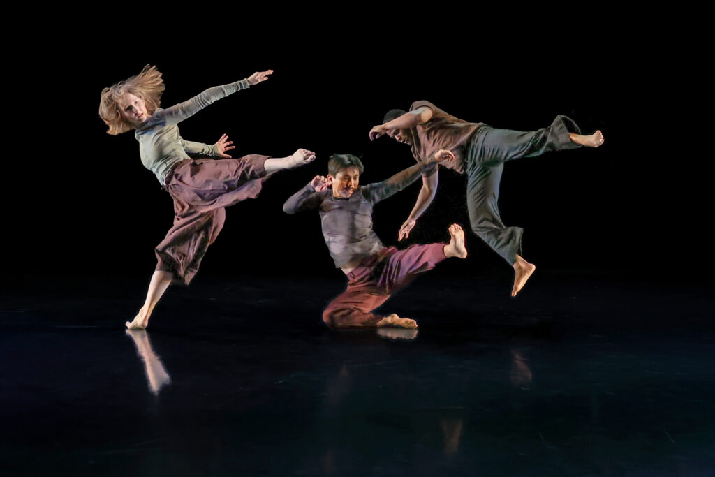 Three dancers in the process of falling on a darkened stage.