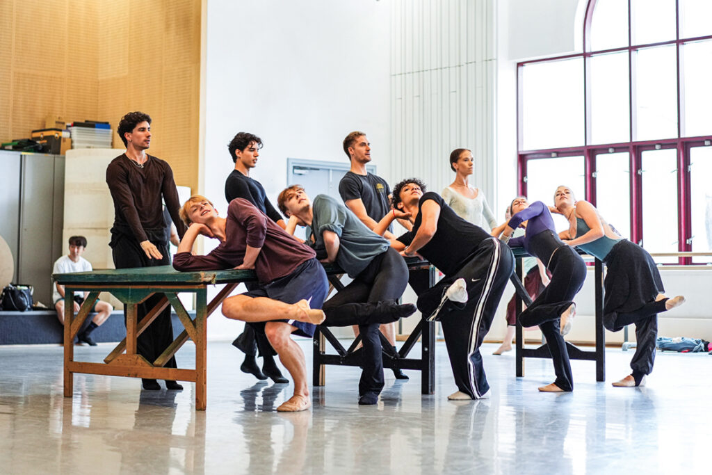 Nine dancers rehearse around a long table. Five lean their upper arms against the table, one leg bending as though sitting with legs crossed. The other four step up to the back of the table in relevé.
