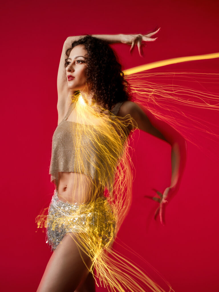 A dancer poses against a red background, golden life arcing around her. She gazes off-camera, arms forming right angles around her head and behind her back.