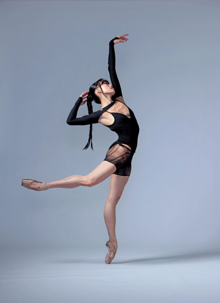 Madeline Woo poses in profile in a low attitude back on pointe. Her upstage arm stretches long overhead, while the downstage arm curves behind her to touch fingertips to the back of her head, mirroring the shape of her working leg in attitude. She wears a black leotard, a pair of ripped black tights that cut off high on her thigh, and a fitted black shrug. Her black hair is in a long braid with strands loose around her face.