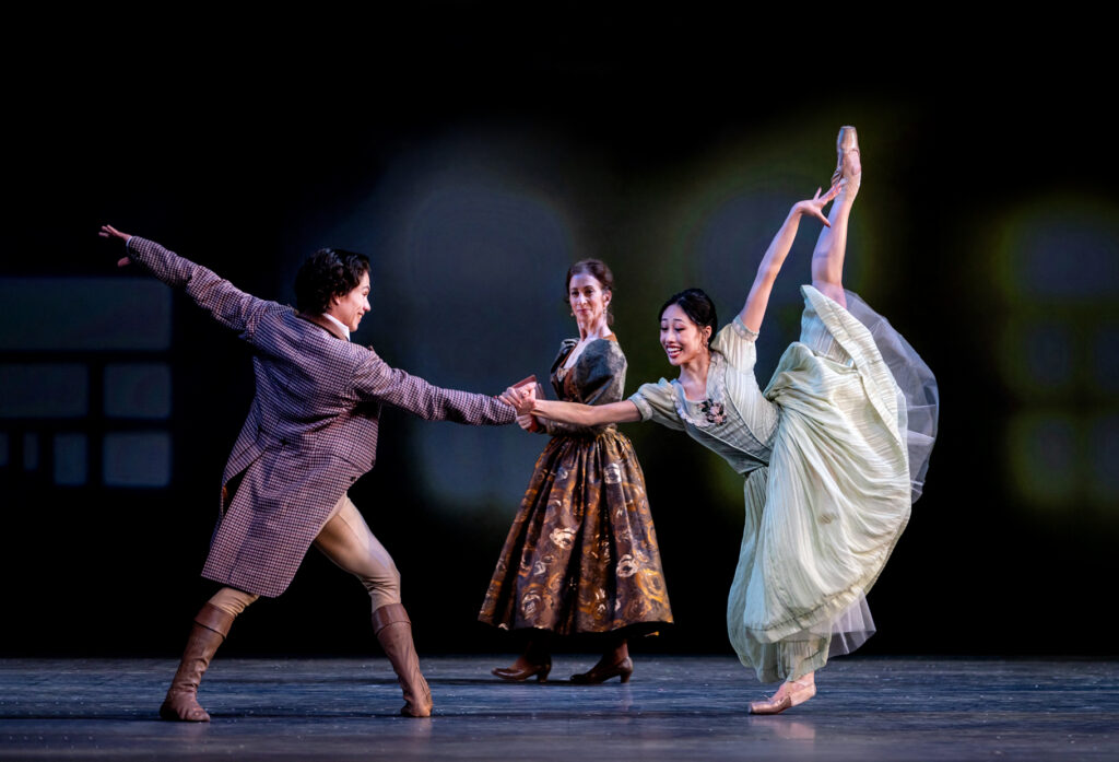 Madeline Woo smiles brightly as she sweeps a leg into a high arabesque penché, a hand outstretched to clasp her partner's, who stands in a fourth position lunge facing her. She is costumed in a pale green dress with a floral pattern at the sternum and pointe shoes. Her partner wears a long, brown check coat over beige tights and brown boots; a woman crossing behind them, looking on with a smile, wears a brown dress and heels.