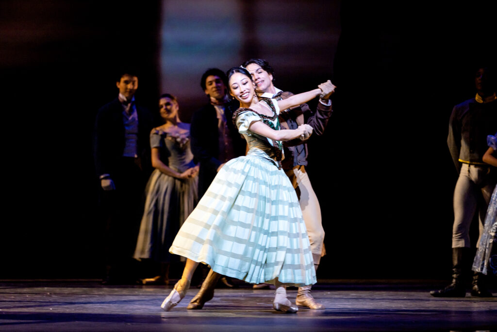 Madeline Woo glances down over her shoulder as she and her partner, hands clasped as they dance side by side, sweep into tendu back with their standing legs in plié. She is costumed in a long dress, with a striped blue skirt and green bodice, and pointe shoes. Couples in similarly period-appropriate costumes linger around the edges of the darkened stage.