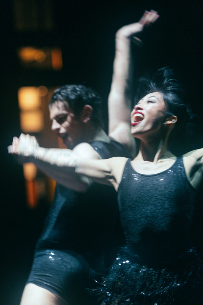 Seen from the wings, Madeline Woo is a blur of motion. She grins widely and joyously as her arms fly wide, one hand clasped with her partner's. She wears a sparkly black tank and beaded skirt.