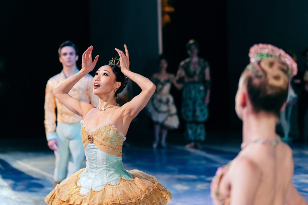 Madeline Woo mimes placing a crown on her head, eyes upturned. She is costumed in a classical tutu in light teal and pale gold.