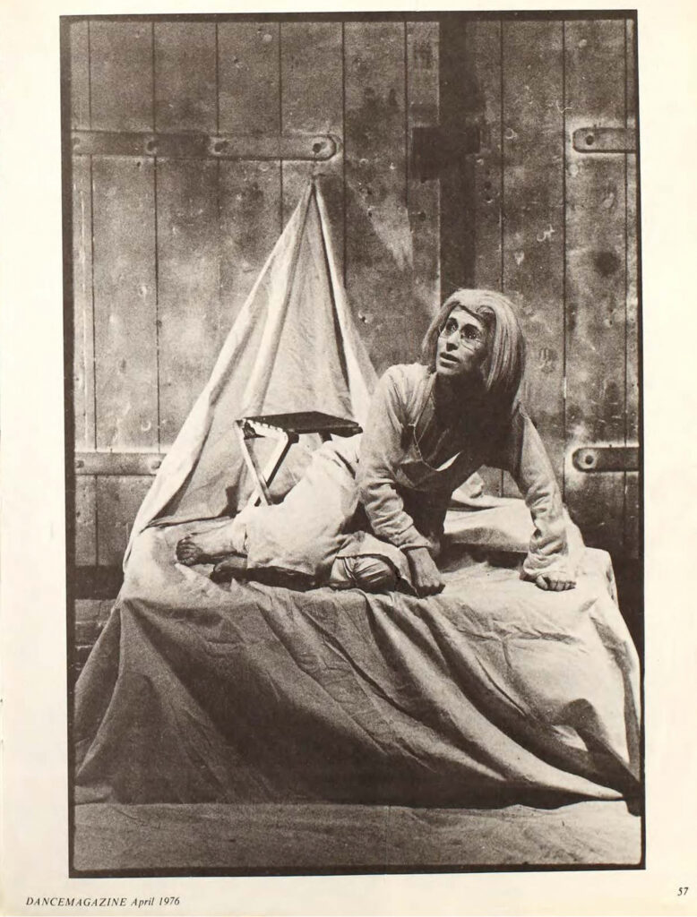 A page from the April 1976 issue of Dance Magazine is mostly filled with a black and white image of Meredith Monk, sitting on one hip with her legs curled beneath her on a bed whose sheets are anchored to the wall behind it and that has a small folding table set just behind Monk. She wears large glasses and what seems to be a wig.