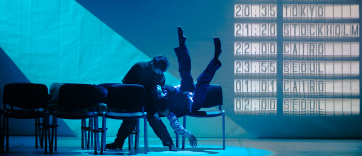 Two dancers sit beside each other in a row of chairs. One has twisted onto their back, legs upraised and akimbo, so their head can rest in the other's lap. The dancer seated upright looks down into their face, knees knocking together. The stage is lit blue, as though moonlight is coming in from a high window; projected on the back wall are departure times for cities like London, Bucharest, Tokyo, and Cairo.