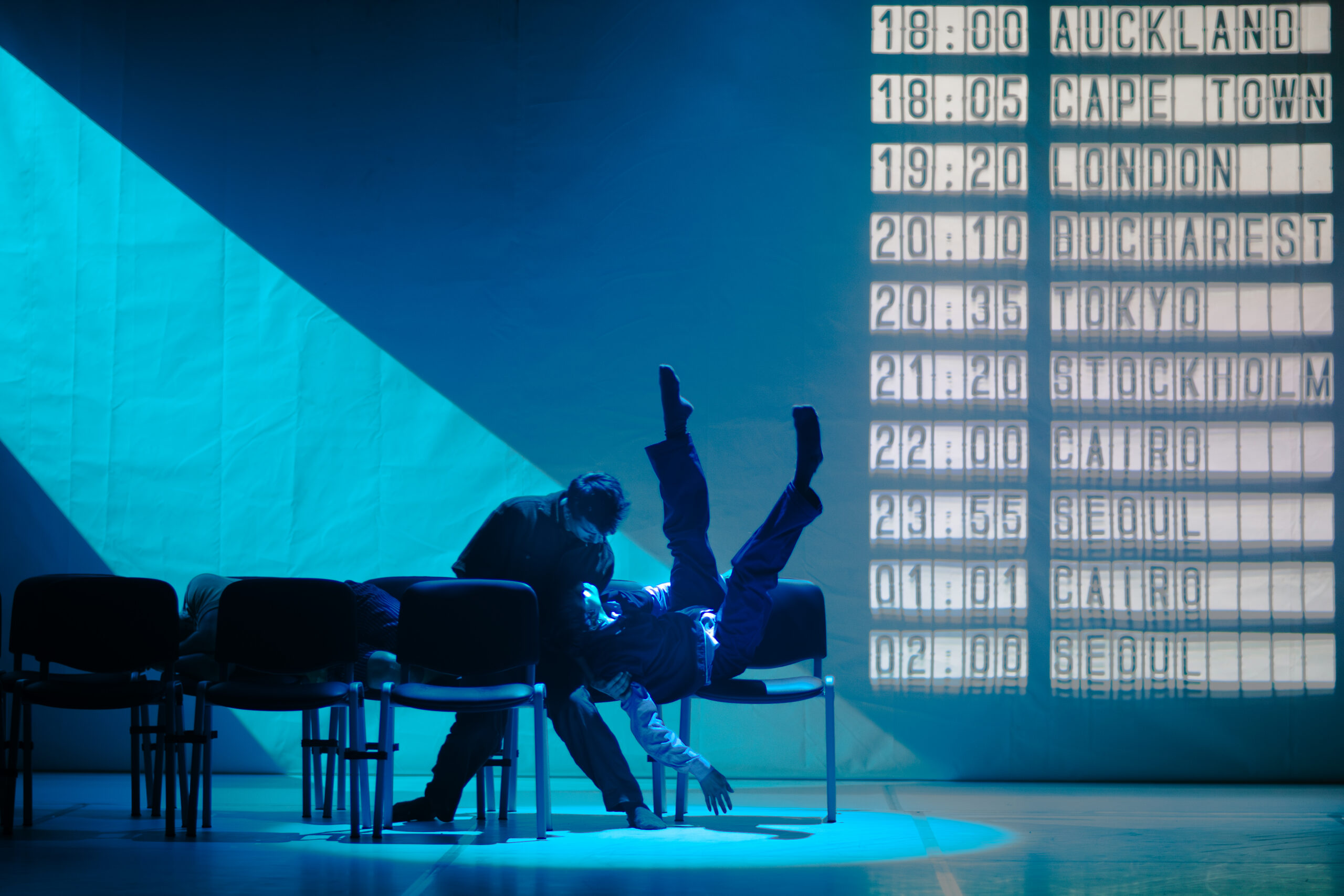 Two dancers sit beside each other in a row of chairs. One has twisted onto their back, legs upraised and akimbo, so their head can rest in the other's lap. The dancer seated upright looks down into their face, knees knocking together. The stage is lit blue, as though moonlight is coming in from a high window; projected on the back wall are departure times for cities like London, Bucharest, Tokyo, and Cairo.