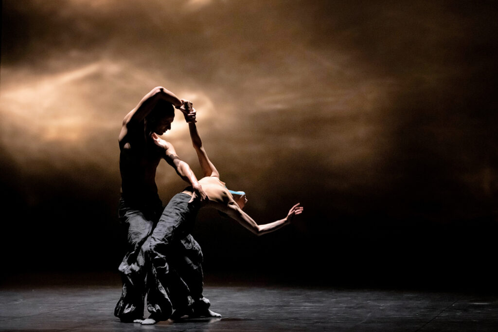 Two identically costumed dancers on a shadowy stage intertwine. One bends back as the other holds them above their hip and by an outstretched arm. They wear baggy black trousers and tight-fitting nude tanks.