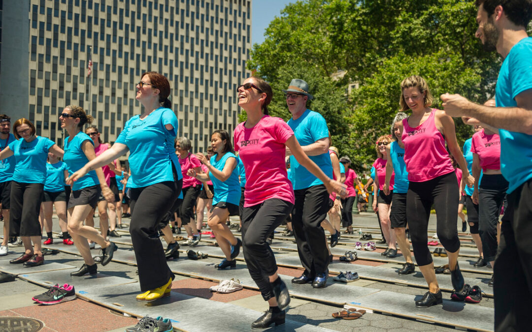 A group of dancers in matching t-shirts and tap shoes perform outside on rows of portable tap boards.