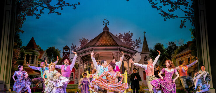 A large ensemble of dancers jump on stage, click their heels, and hike their skirts. The women wear patterned smocked dresses and the men vests and bow ties.