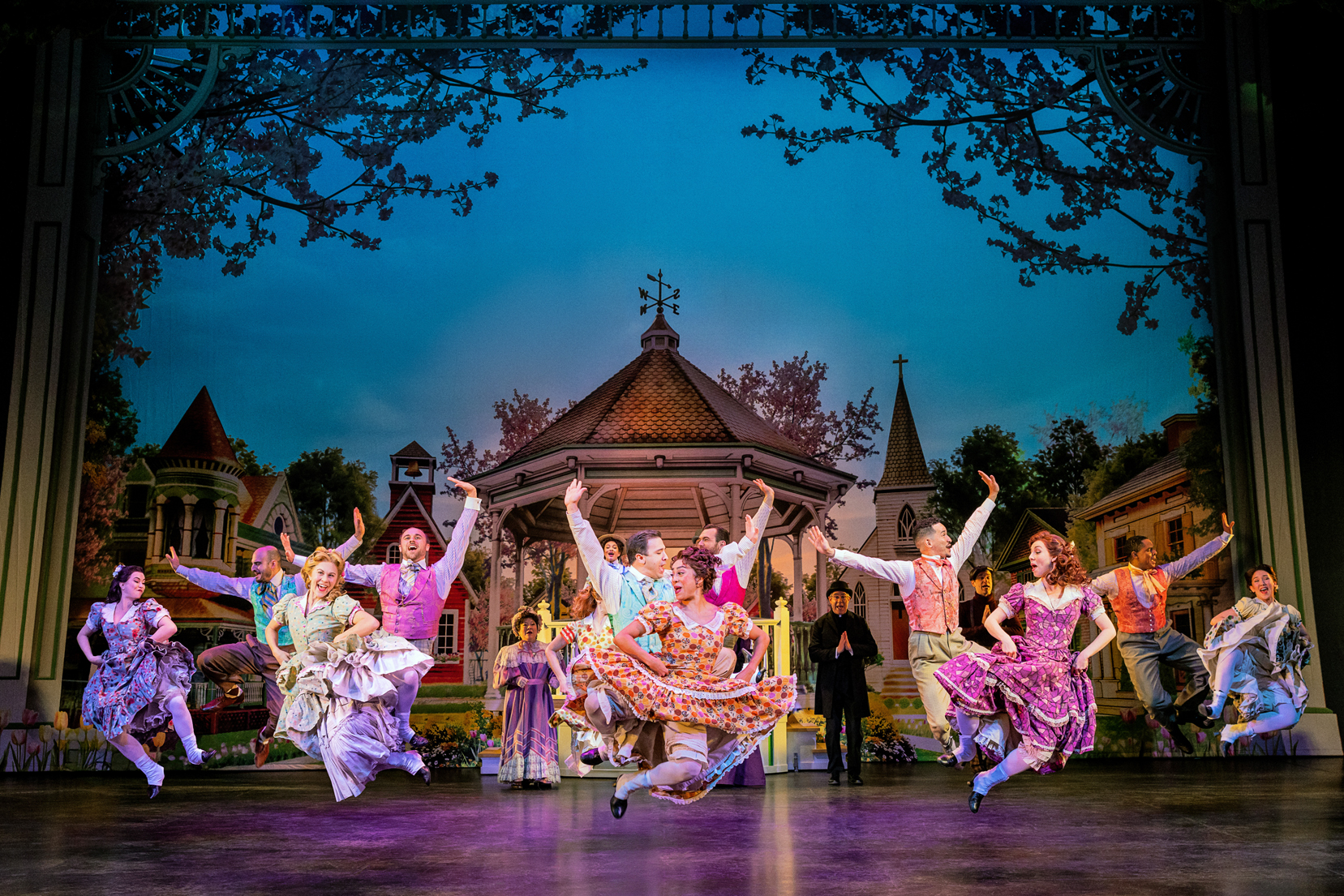 A large ensemble of dancers jump on stage, click their heels, and hike their skirts. The women wear patterned smocked dresses and the men vests and bow ties.