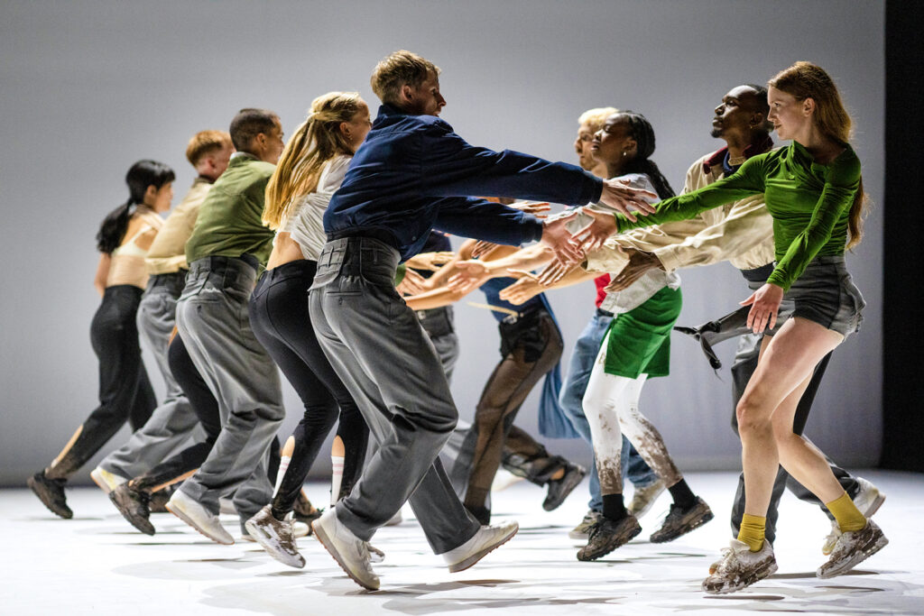 A line of two dancers faces each other, arms outstretched to meet their partners as they step forward. They are costumed in streetwear; many have mud visibly splattered on their shoes and lower legs.