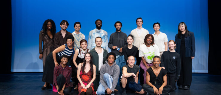A large group of dancers stands on a stage in front of a blue background, all smiling at the camera.