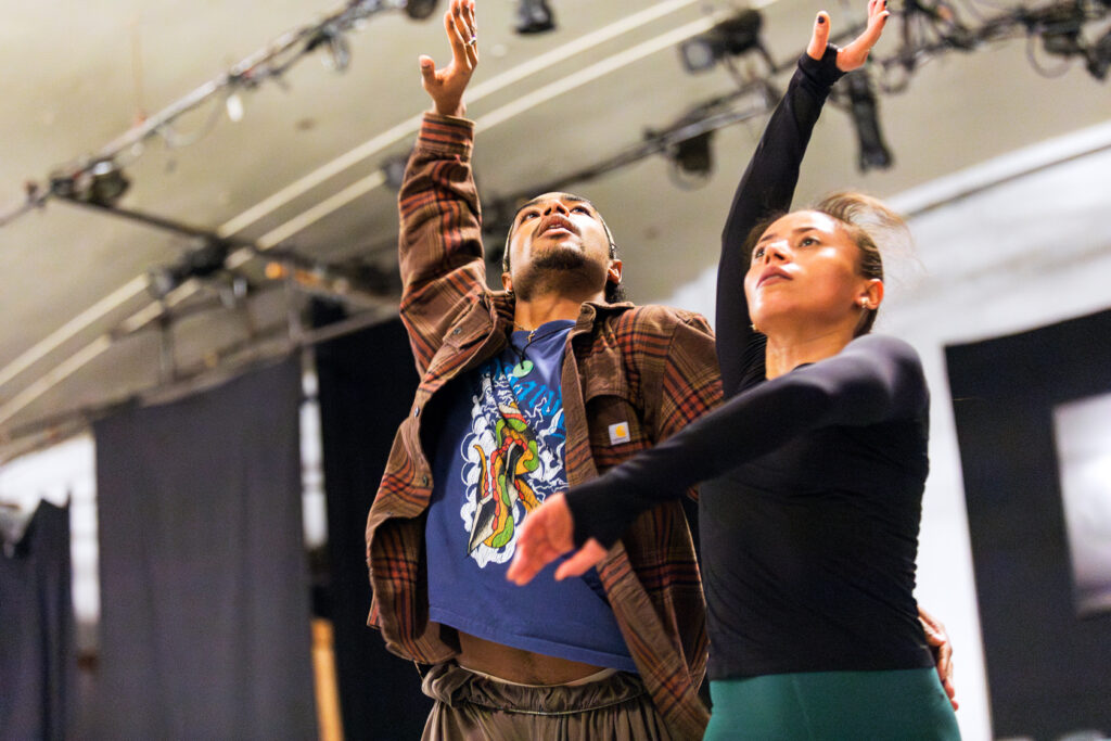 Two dancers in rehearsal are shown from the waist up. They arch back slightly, eyes raised, as they each bring one arm overhead.
