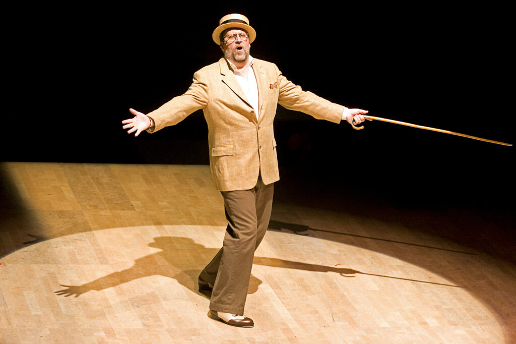 Tony Waag performs in a spotlight on a wooden floor, arms open to the audience with a hook-handled cane in one hand.