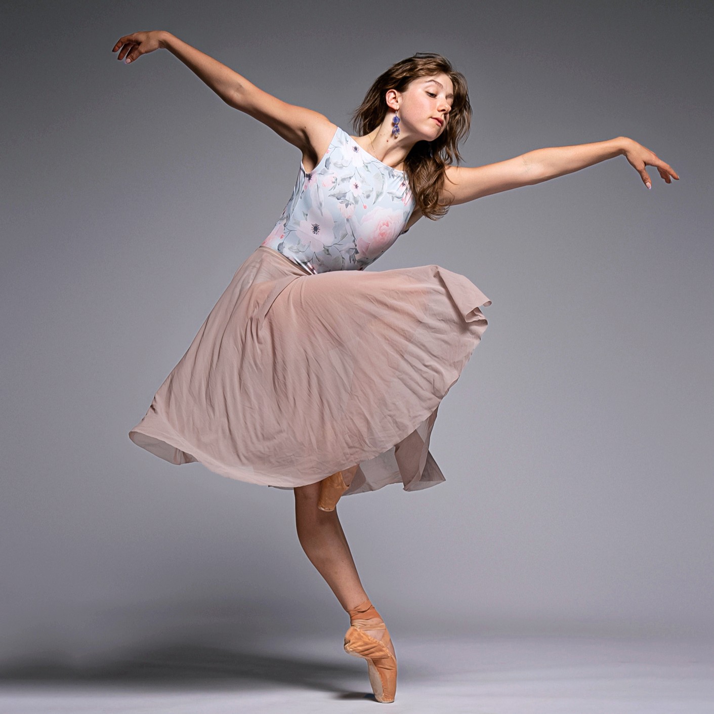 Aurora Chernis wears a floral leotard, knee-length pink skirt, and earrings. She poses en pointe with her right leg in parallel passe.