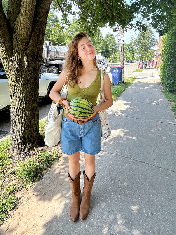 Aurora Chernis wearing a tank top, jean shorts, and cowboy boots while holding a small watermelon. 