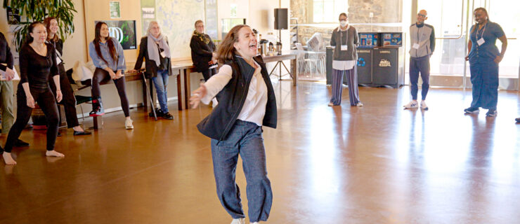 Ferro-Murray dances across the floor in a room with our artists. They wear pedestrian clothing.