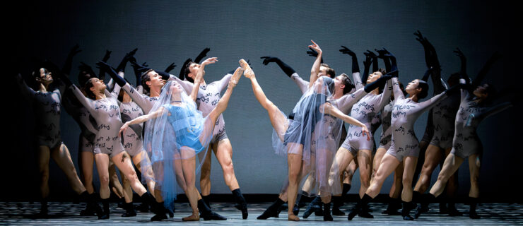 Corps dancers face each other onstage while extending their upstage leg in tendu devant with their arms lifted in a V overhead. Two lead dancers stand in the middle performing a developpe devant. Their costumes include mesh fabric, unitards, and black gloves.