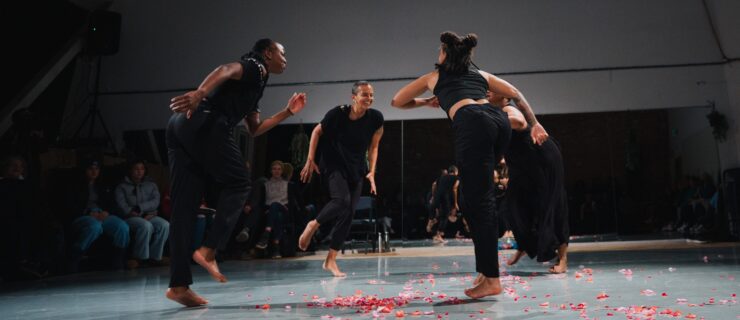 Four dancers costumed in black face each other in a circle. They smile joyfully as they cant toward the center, knees raised mid-step. Red flower petals are scattered across the studio floor. Seated audience members are dimly lit at the edges of the space.