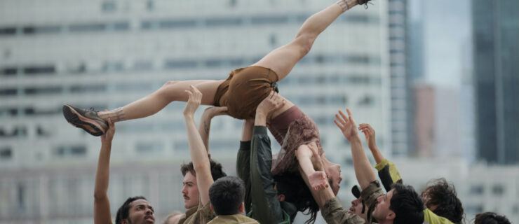 A group of dancers in earth-toned street clothes performs outdoors in an urban setting. One woman is raised dramatically above the crowd in an upside-down split lift.