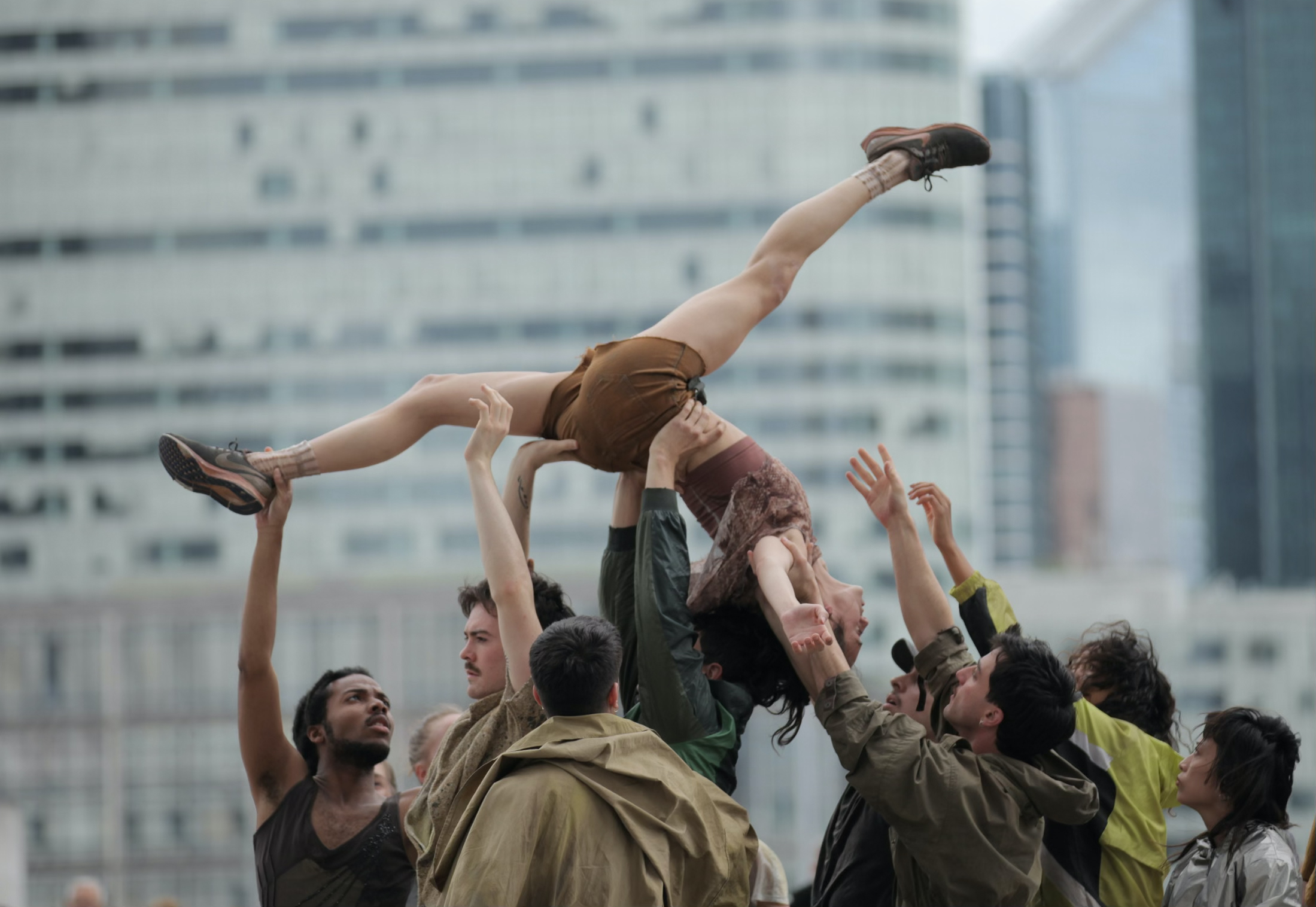 A group of dancers in earth-toned street clothes performs outdoors in an urban setting. One woman is raised dramatically above the crowd in an upside-down split lift.