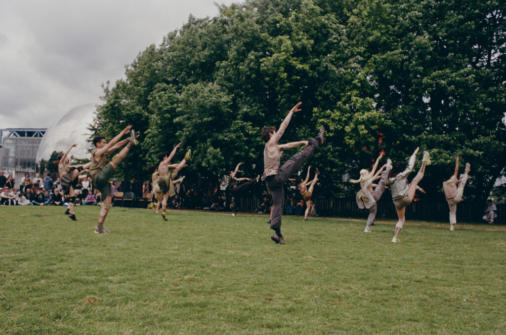 A large group of dancers in earth-toned street clothes and sneakers performs outdoors on a large lawn. They kick their right legs in front of them and reach their arms toward their working legs.