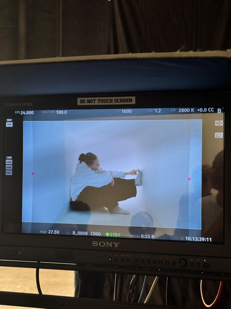 A camera point of view of a performer sitting in a white box with one foot pressing against the wall.