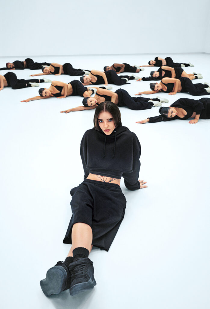 Young Miko sits on the floor, resting back on her hands. A group of dancers dressed in black lie on their sides behind her. They film in an empty white production studio.