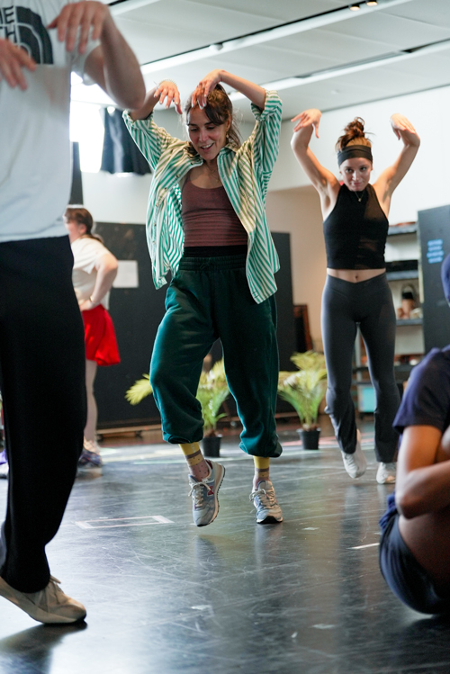 In a rehearsal studio, Delgado—wearing a green striped button-down shirt and dark-green sweatpants—moves with a group of dancers, her arms raised, a smile playing on her face.