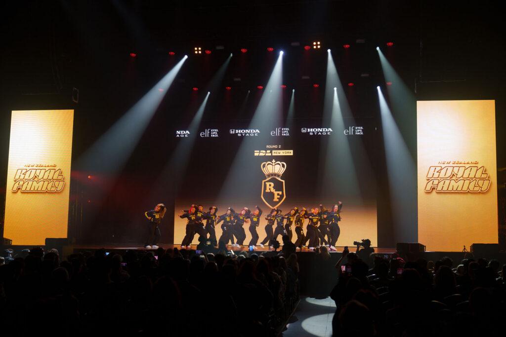 A large group of dancers wearing black-and-yellow jerseys and pants performs on a stage dramatically lit in gold. The Royal Family logo, topped with a crowd, is projected on a screen at the rear of the stage.