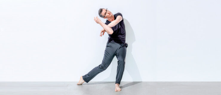 Sean Dorsey dances in front of a white backdrop. He leans to the side as he drags the top of a pointed foot against the floor, knees soft. His arms bend so a wrist rests against the opposite forearm, the angular shape pulling to the side as his head tips over, following gravity's pull. His short gray hair is styled in a faux-hawk; his expression, behind cat-eyed glasses, is sharp. He wears a dark t-shirt and grey pants.