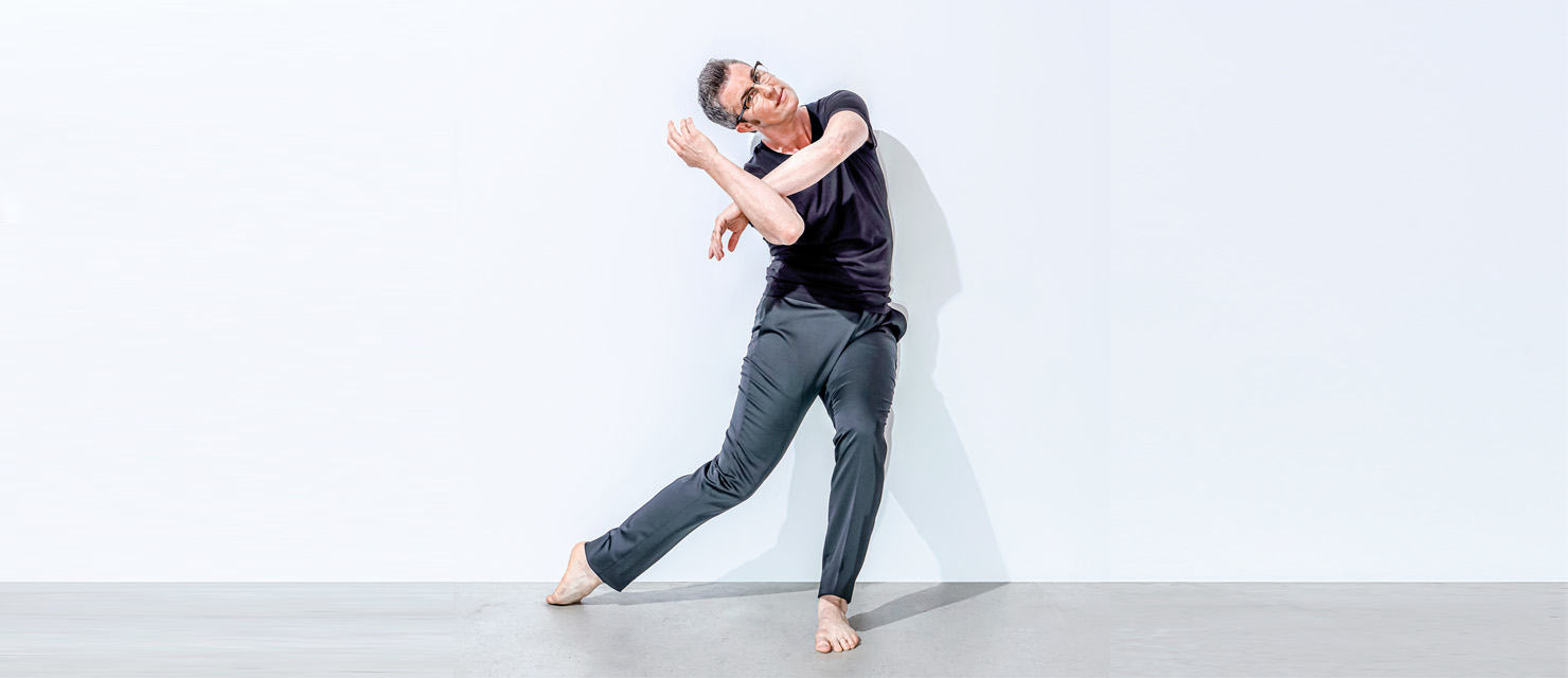 Sean Dorsey dances in front of a white backdrop. He leans to the side as he drags the top of a pointed foot against the floor, knees soft. His arms bend so a wrist rests against the opposite forearm, the angular shape pulling to the side as his head tips over, following gravity's pull. His short gray hair is styled in a faux-hawk; his expression, behind cat-eyed glasses, is sharp. He wears a dark t-shirt and grey pants.