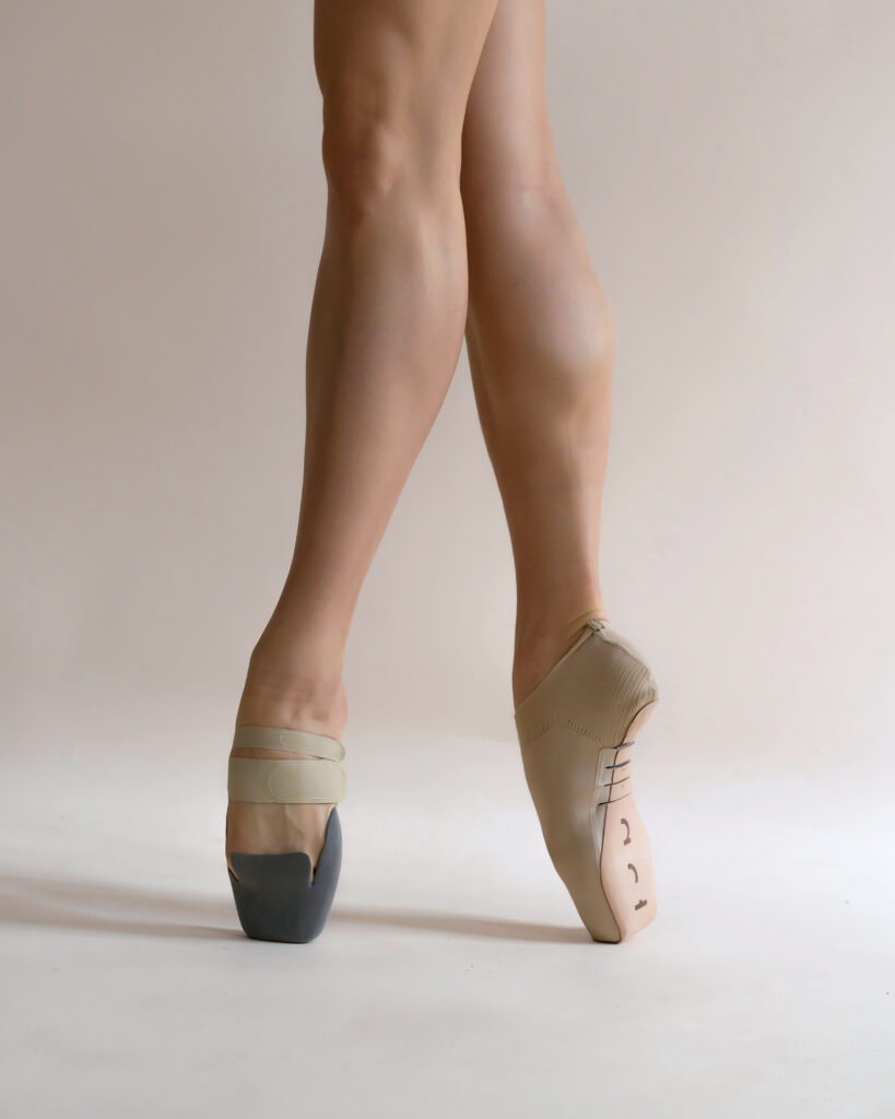 A dancer wears a 3-D printed shoe on one foot and a knitted tan pointe shoe on the other while standing in 4th position.