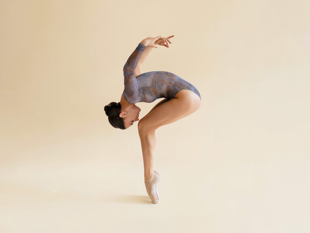 A dancer wearing a three quarter purple and brown leotard contracts over en pointe with their arms swinging backwards.