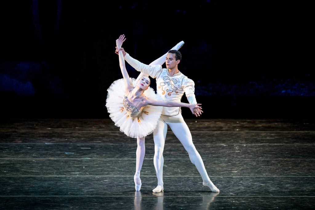 Mira Nadon balances in a partnered back attitude penché, wrists held by Ryan Tomash. She wears a white classical tutu, pink tights, and pointe shoes; Tomash is in a matching long-sleeved tunic, white tights, and slippers.